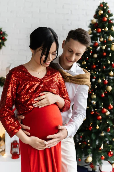 positive man touching belly of pregnant asian wife in festive clothes near Christmas tree on blurred background