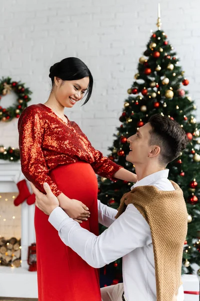 若い男と妊娠中のアジア人の妻がクリスマスツリーの近くで笑顔でお祝いの服を着て — ストック写真