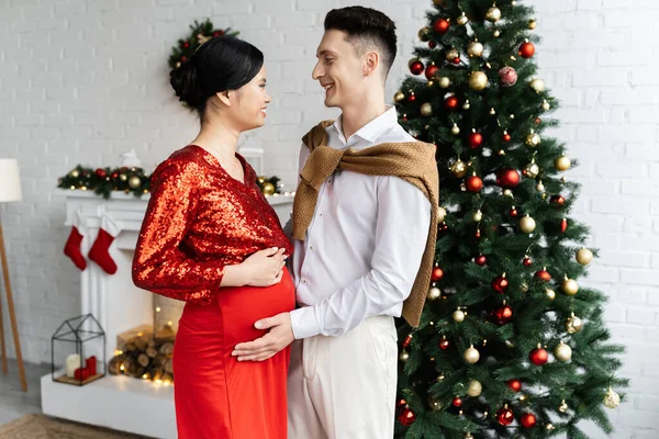 妊娠中の優雅なアジア系女性と夫の笑顔がクリスマスツリーの近くのリビングルームで — ストック写真