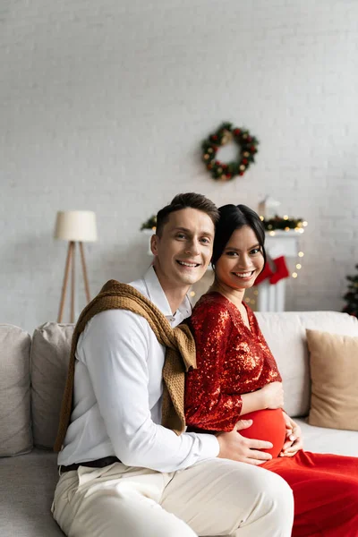 Cheerful Man Pregnant Asian Wife Festive Outfit Looking Camera Couch — Stock Photo, Image