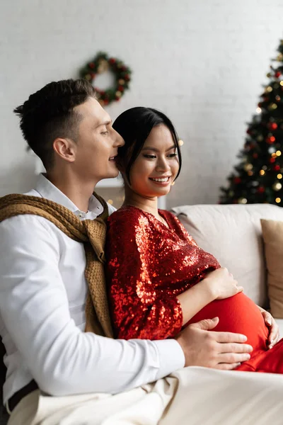 妊娠中でエレガントなアジアの女性は夫の近くで笑みを浮かべて彼女のおなかを抱きますクリスマスの装飾付きのリビングルームで — ストック写真