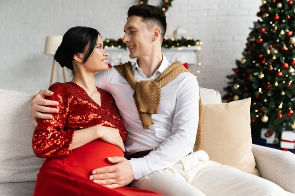 妊娠中のアジアの女性と夫のお祝いの服でお互いを見ます上のソファの近くにぼやけたクリスマスツリー — ストック写真