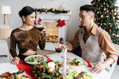 Mutlu Asyalı kadın romantik bir akşam yemeğinde şarap kadehi tutuyor kocasıyla birlikte bulanık Noel ağacının yanında.