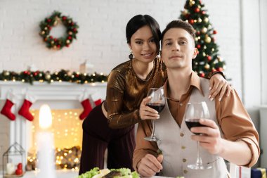 Neşeli Asyalı kadın elinde şarap kadehiyle ve Noel yemeğinin yanında kocasını kucaklıyor. 