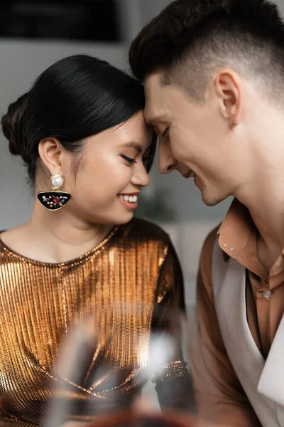 happy asian woman in shiny blouse smiling face to face with young husband on blurred foreground