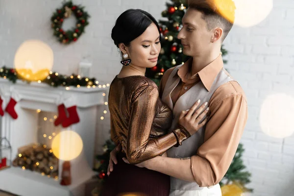 雅致的跨种族夫妇在客厅里拥抱 靠近模糊的壁炉 装饰着圣诞装潢 — 图库照片