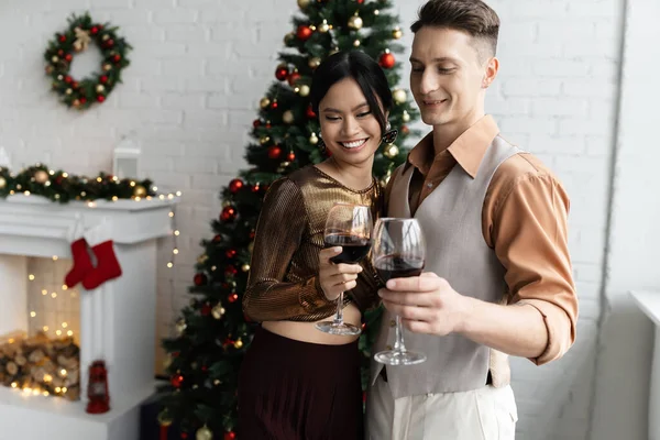 笑着的多民族夫妇在圣诞树旁举杯红葡萄酒 — 图库照片