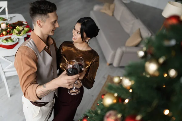 在圣诞树附近 穿着时髦服装的快乐的跨种族夫妇们的高视角的酒杯碰杯 — 图库照片
