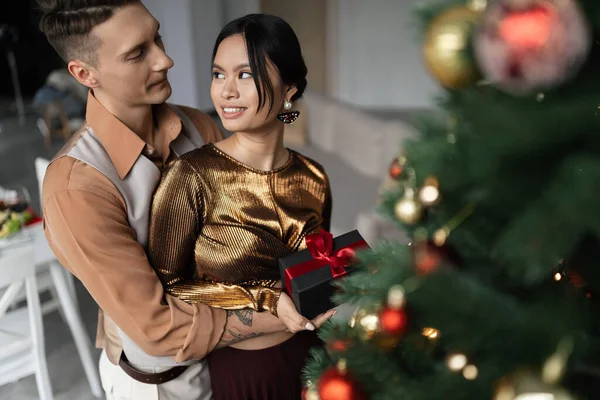 Tattooed Man Hugging Happy Asian Wife While Holding Christmas Present — Stock Photo, Image