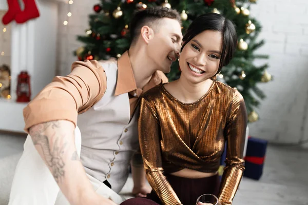 快乐的男人坐在圣诞树下 在快乐的亚洲妻子的耳边窃窃私语 — 图库照片