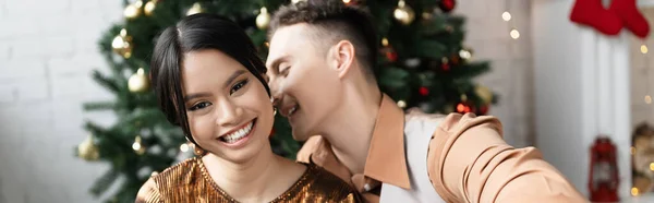 cheerful man whispering in ear of happy asian wife near Christmas tree, banner