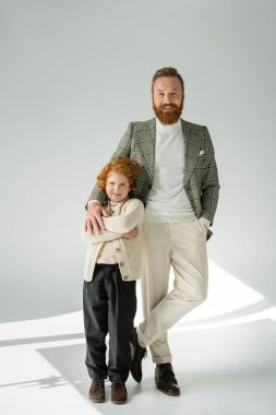 Full length of stylish father and redhead son posing and looking at camera on grey background with light  clipart