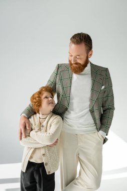 Stylish redhead boy looking at bearded dad in checkered jacket on grey background with sunlight  clipart