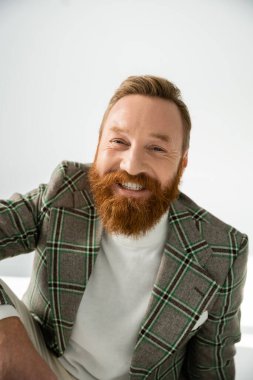 Ekose ceketli sakallı adam gri arka planda kameraya bakıyor.