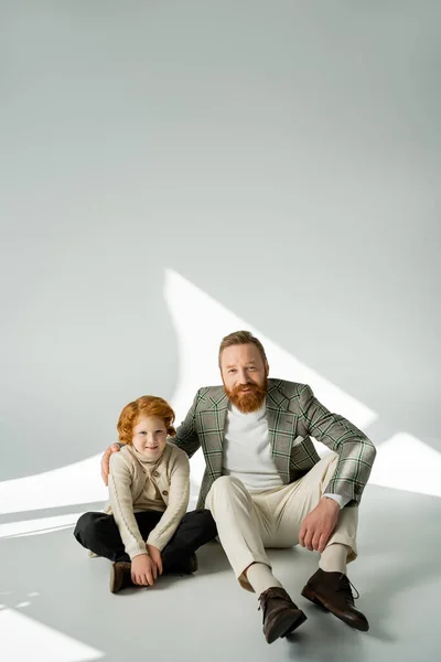 Positive red haired father and son looking at camera while sitting on grey background with light