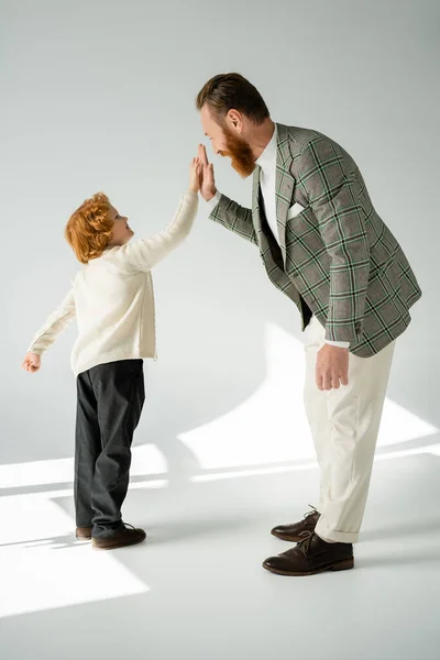 Side view of positive and stylish man giving high five to red haired son on grey background