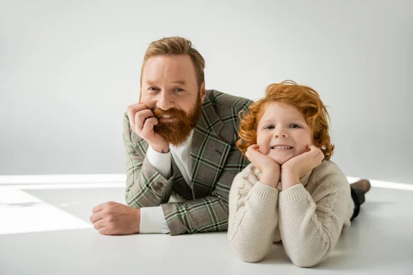 cheerful redhead father and son in stylish clothes lying and looking at camera on grey background