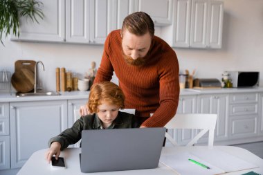 Sakallı adam kızıl saçlı oğlana laptopun yanında ödev yaptığını anlatıyor.