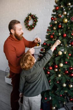 Kızıl saçlı baba ve oğul ellerinde mücevherlerle evde çam ağacı süslüyorlar.