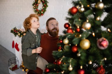 Yüksek açılı, sakallı bir adamın kızıl saçlı bir oğlanı, bulanık ön planda süslü bir Noel ağacının yanında tutuşu.