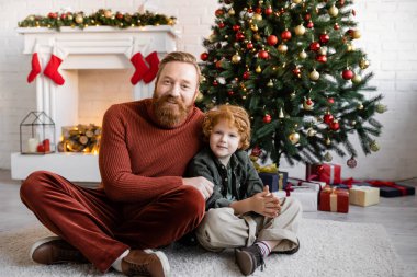 Kızıl saçlı baba ve oğul oturma odasında bacak bacak üstüne atmış Noel ağacı ve dekore edilmiş şömineyle oturuyorlar.