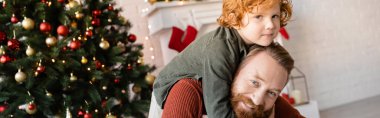happy bearded man piggybacking redhead son near blurred Christmas tree, banner clipart