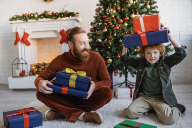 Çocuk hediye kutularını kafasının üstünde tutarken sakallı babası Noel ağacının yanında oturmuş eğleniyor.