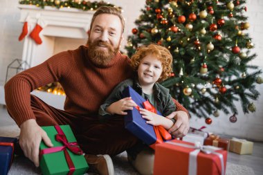 Sakallı babasıyla gülümseyen kızıl saçlı çocuk arka plandaki hediye kutularının yanındaki kameraya ve Noel ağacına bakıyor.
