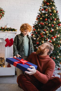Hayret bir adam elinde Noel hediyesiyle gülümseyen kızıl saçlı bir oğlan ve süslü bir çam ağacı tutuyor.