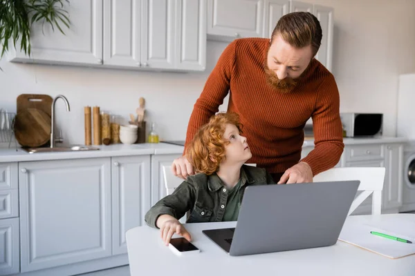 bearded man talking to redhead son learning near notebook and smartphone with blank screen in kitchen