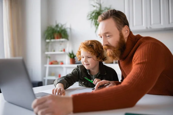 红头发的父亲和儿子在一起做作业的时候看着模糊的笔记本电脑 — 图库照片
