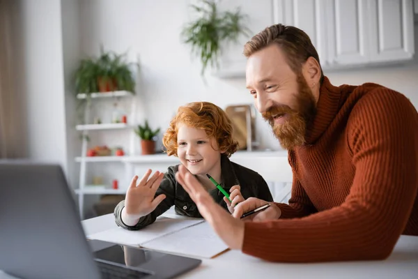 一緒に宿題をしながらノートパソコンの近くで手を振って幸せな赤毛の父と息子 — ストック写真
