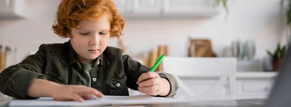 红头发的孩子拿着笔在模糊的笔记本边 一边在厨房做家庭作业 一面横幅 — 图库照片