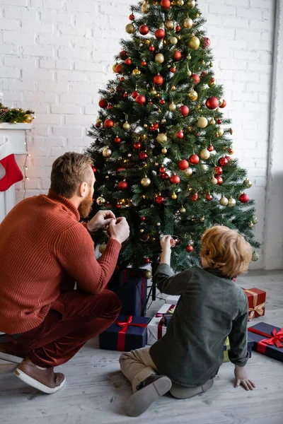 ギフトボックスの近くにクリスマスツリーを飾る赤毛の父と息子のバックビュー — ストック写真