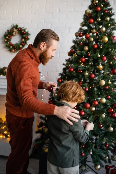 家でクリスマスツリーを飾るレッドヘッドの息子の近くに立つ髭の男 — ストック写真