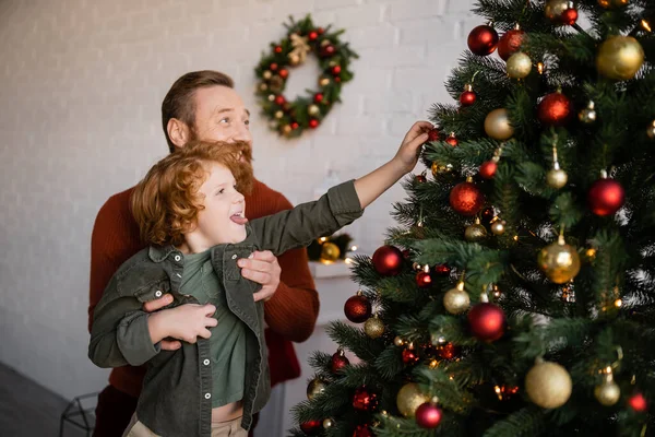 男人牵着红头发的儿子伸出舌头 用圣诞彩礼装饰松树 — 图库照片