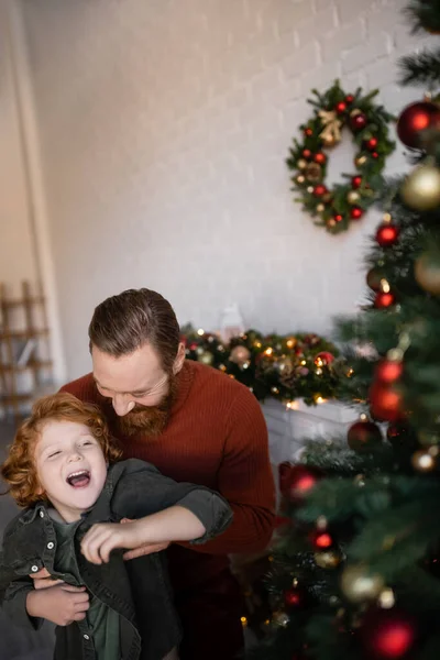 ぼやけたクリスマスの装飾の近くで笑っている赤毛の息子と遊ぶ男 — ストック写真