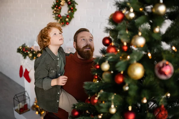 大胡子男子将红头发的儿子抱在装饰过的圣诞树旁的高角镜模糊的前景 — 图库照片