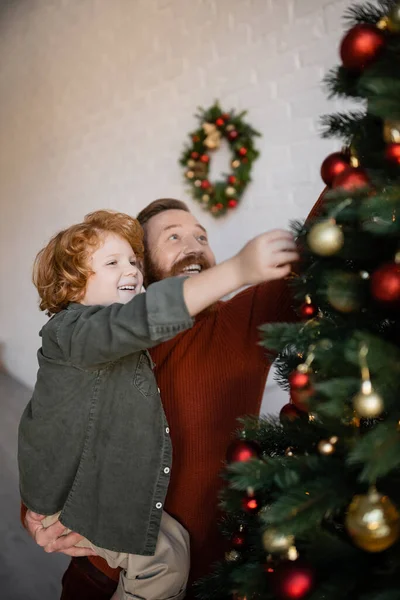 happy man with redhead son decorating Christmas tree on blurred foreground