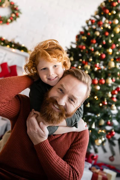 joyful father and son with red hair looking at camera while having fun near blurred Christmas tree