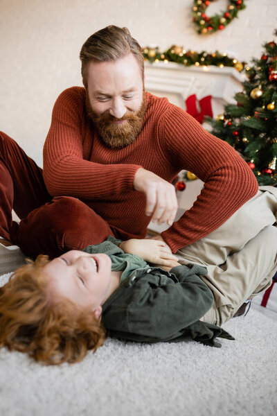 happy bearded man playing with laughing son lying on floor in living room with Christmas decor