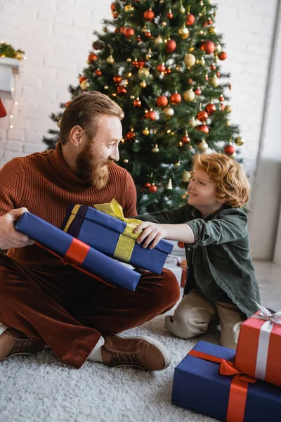 Mutlu kızıl saçlı baba ve oğul birbirlerine hediye kutularının ve Noel ağacının yanında bakıyorlar.