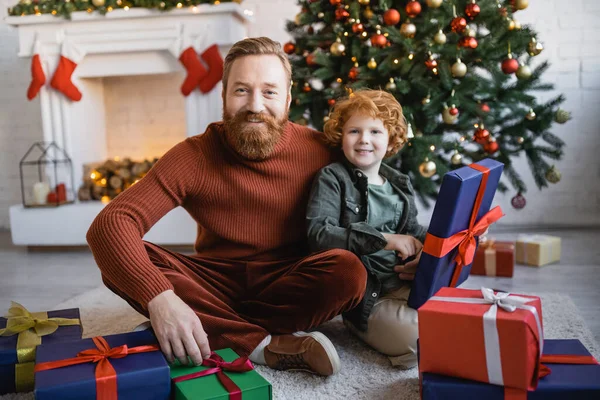 bearded man with redhead kid smiling at camera while sitting on floor near gift boxes and decorated Christmas tree