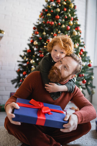 Рыжий ребенок обнимает счастливого бородатого папу, сидящего на полу с рождественским подарком