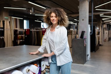 cheerful and curly saleswoman standing near rack with different fabric rolls in textile shop  clipart