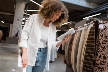 happy saleswoman using digital tablet near patterned fabric in textile shop  clipart