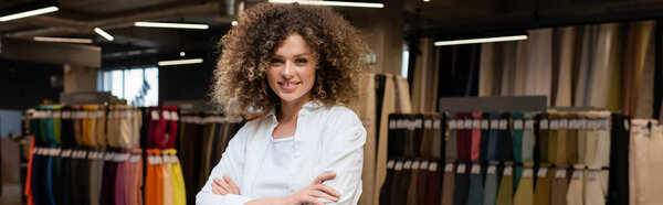 cheerful and curly saleswoman standing with crossed arms in textile shop, banner