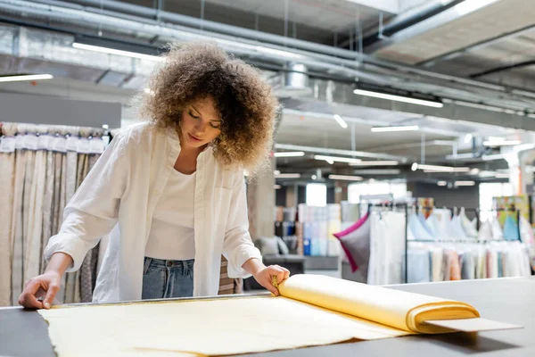 stock image young and curly saleswoman measuring yellow linen fabric roll in textile shop 