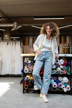 full length of cheerful salesperson with curly hair standing with hand in pocket near rack with fabric rolls   clipart