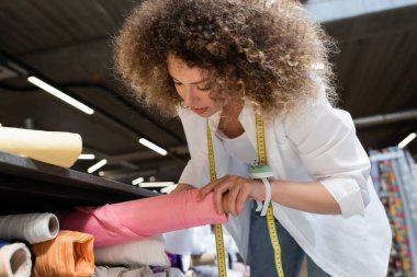 Kıvırcık satıcı kadın tekstil dükkanındaki raftan pembe kumaş rulo alıyor.